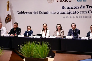 Siete funcionarios sentados a una mesa de presídium con micrófonos, botellas de agua y carteles de identificación delante de una mampara