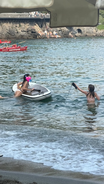 Dos niñas en el mar, una sobre una tabla de paddle surf inflable, la otra sujetando una cuerda. Al fondo, costa rocosa con personas y botes salvavidas
