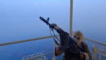 Imagen de una persona enmascarada sosteniendo un rifle negro, de pie sobre una estructura metálica con el vasto mar azul y nublado como fondo