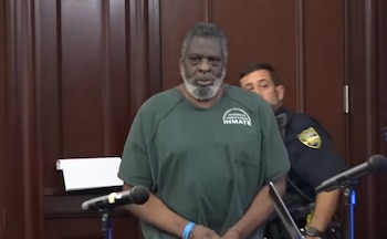 Hombre afroamericano de edad, con barba gris, vistiendo uniforme de recluso verde y pulsera azul, en sala con fondo de madera. Se ve un oficial de policía y micrófonos
