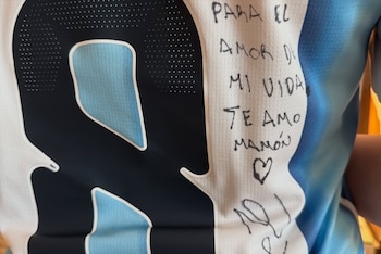Primer plano de una camiseta de fútbol blanca y celeste con un gran número oscuro. Contiene un mensaje manuscrito en tinta negra, un dibujo de corazón y una firma