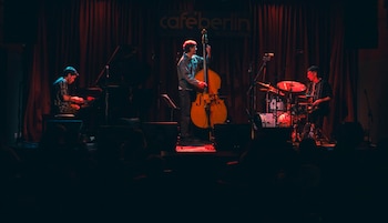Un trío musical toca en un escenario con cortinas rojas oscuras y el letrero "caféberlin Buenos Aires". Un pianista, un contrabajista y un baterista actúan