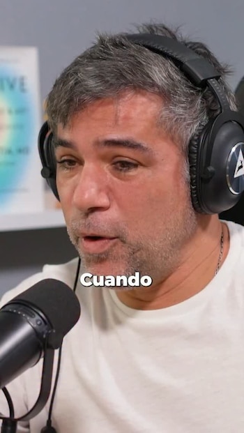 Hombre de mediana edad con cabello gris y barba, vestido con camiseta blanca, usando auriculares negros y hablando en un micrófono, con la palabra 'Cuando'