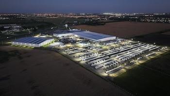 Vista aérea nocturna de un complejo industrial iluminado con grandes naves y cientos de camiones estacionados en hileras junto a una carretera