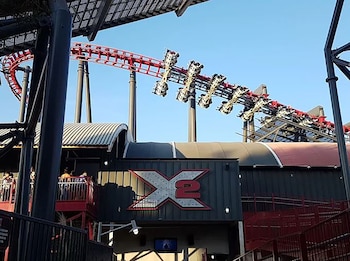 Una vía de montaña rusa de color rojo sobre soportes oscuros con un tren en marcha y una fachada metálica con el letrero X2