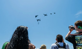 Un grupo de personas, vistas desde atrás, observan hacia un cielo azul brillante donde varias aeronaves militares, incluyendo un B-2, B-1B y B-52, vuelan en formación