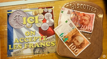 Un cartel con monedas francesas y la frase 'ICI ON ACCEPTE LES FRANCS' junto a billetes de 100 y 200 francos sobre un objeto marrón con 'BISCOTTES' grabado