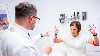 Un médico en bata blanca de espaldas apunta a las manos de una paciente sentada en una camilla de examen, con equipo médico en la pared