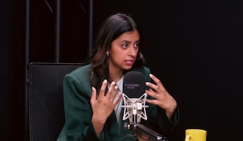 La Dra. Trisha Pasricha, con cabello oscuro y blazer verde, gesticula con las manos frente a un micrófono de podcast en un estudio oscuro