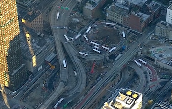 Vista aérea de rampas viales elevadas con autobuses en tránsito, maquinaria de construcción en el terreno y edificios circundantes