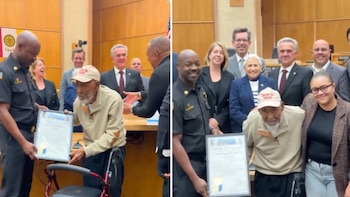 Un bombero entrega un certificado a anciano con gorra y barba blanca, junto a andador, rodeado de sonrientes personas en una sala pública
