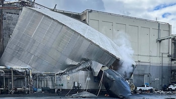 Vista de un edificio industrial parcialmente colapsado, con una gran estructura metálica inclinada, vapor blanco saliendo y escombros dispersos en el suelo