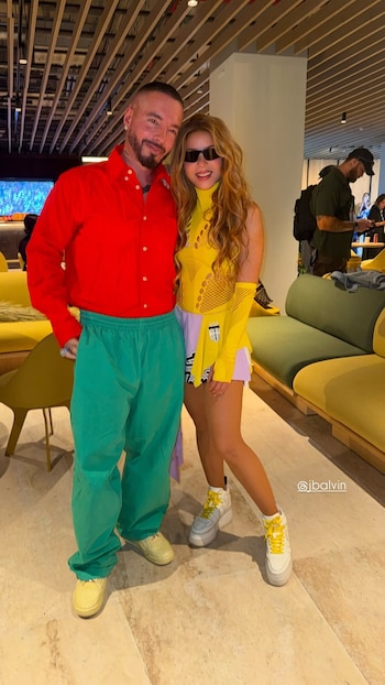 Fotografía de Shakira y J Balvin de pie, sonriendo y mirando a cámara. J Balvin lleva camisa roja y pantalón verde; Shakira, top amarillo, falda lila y gafas de sol