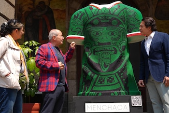 Tres personas observan una escultura gigante de un torso verde con diseños prehispánicos y bandas tricolores sobre una base con el nombre 'Menchaca'