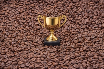Una copa dorada sobre una base negra, llena de granos de café, se encuentra en el centro de una superficie cubierta por más granos de café tostado.