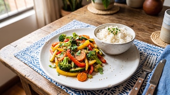 Un plato blanco con brócoli, pimientos y zanahorias salteadas junto a un tazón con arroz blanco sobre un mantel azul.