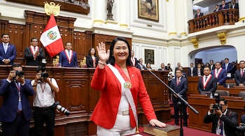 Keiko Fujimori con saco rojo y banda presidencial, saluda con mano levantada en el Congreso de Perú. Un micrófono, un libro y fotógrafos la rodean.