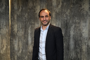 Un hombre con barba corta, camisa clara y saco oscuro delante de una pared gris con una línea de luz vertical