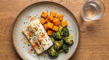 Un plato con un filete de merluza, cubos de calabaza asados y ramilletes de brócoli asados. Al lado hay un vaso de agua sobre una mesa de madera.