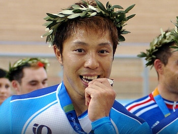 Primer plano de un hombre sonriente mordiendo una medalla con inscripción "ATHENS 2004". Lleva una corona de laurel y uniforme deportivo azul