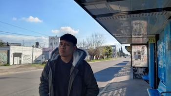 Un joven con chaqueta espera en una parada de autobús con cartel de Línea 176. Se aprecian una calle, una acera, edificios y cielo azul con nubes