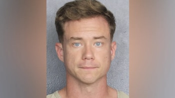 Primer plano de un hombre con pelo castaño claro y ojos azules, mirando directamente a la cámara sobre un fondo gris liso, similar a una ficha policial