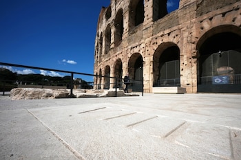 Vista del Coliseo de Roma con sus arcos de piedra y el cielo azul claro. Un hombre de traje camina en el medio, con suelo pavimentado en primer plano
