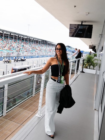 Antonela Roccuzzo, con lentes de sol y vestida de negro y pantalones anchos claros, sonríe apoyada en una barandilla, observando la pista del Gran Premio de Miami