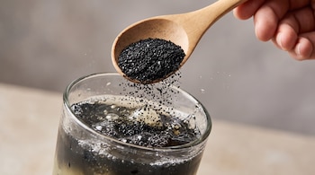 Mano sujetando cuchara de madera que vierte carbón activado granulado en un vaso con limonada negra, burbujas y salpicaduras en la superficie.
