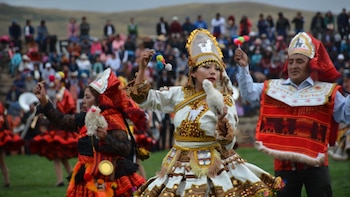 Varias personas con trajes folclóricos bordados y tocados tradicionales bailan en un campo frente a una multitud