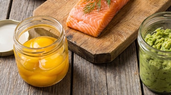 Un frasco de vidrio con yemas de huevo, un filete de salmón crudo con eneldo en una tabla de madera y un frasco con puré de palta sobre una superficie de madera.