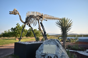 Réplica de esqueleto de dinosaurio de cuerpo completo en un parque. Un cartel rocoso, una palma y árboles verdes se ven bajo un cielo azul claro con montañas de fondo