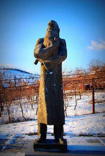 Escultura de un hombre barbudo, vestido con túnica y brazos cruzados, en un paisaje invernal con nieve, árboles desnudos y colinas, bajo un cielo azul