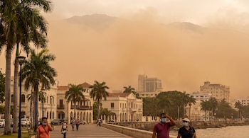 Un paisaje urbano costero con palmeras, edificios y montañas cubiertas por una densa capa de polvo, personas caminan en primer plano usando mascarillas.