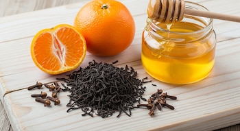 Té negro suelto, una mandarina entera y otra partida por la mitad, clavos de olor y un frasco de miel con un cucharón de madera sobre una tabla clara.