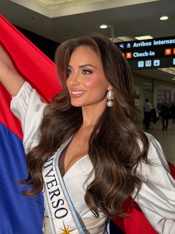 Tamara Rogouski, de perfil y sonriendo, con un vestido blanco, banda de Miss Universo, aretes de perla y una bandera roja y azul