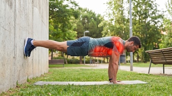 Un hombre en ropa deportiva realiza una plancha con los pies apoyados en una pared de concreto, sobre una colchoneta en un parque con césped verde.