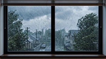 Ventana con gotas de agua sobre el vidrio que muestra casas, árboles sacudidos por el viento y cielo gris nublado