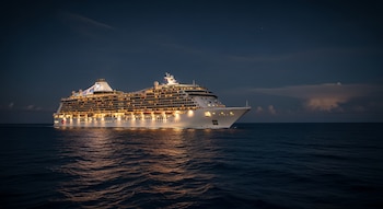 Vista de un gran crucero blanco brillantemente iluminado navegando en un mar oscuro bajo un cielo nocturno profundo, con reflejos dorados en el agua.