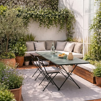 Patio exterior luminoso con paredes cubiertas de plantas. Mesa verde plegable con sillas, botella de agua y aperitivos. Un sofá con cojines y plantas en macetas.