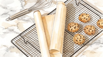 Un batidor de varillas, dos rollos de papel de horno y cinco galletas con chispas de chocolate sobre una rejilla de enfriamiento en una superficie marmolada.