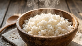 Primer plano de un cuenco de madera lleno de arroz blanco cocido, del que se eleva vapor. Una cuchara de madera y arroz crudo están en la mesa.