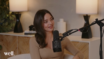Miranda Kerr, con cabello castaño, usa una blusa beige y sonríe mientras habla por un micrófono de estudio negro frente a una estantería con lámparas y libros