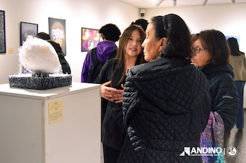 Una joven habla con dos mujeres junto a una escultura sobre un pedestal blanco, dentro de una sala con pinturas en la pared
