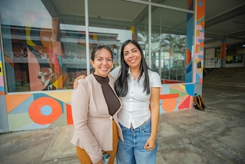 Dos mujeres sonrientes posan de pie, una con cabello largo suelto y la otra recogido, frente a un edificio con ventanas y un mural abstracto colorido