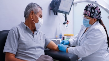 Una profesional con bata blanca, guantes y mascarilla extrae sangre del brazo de un hombre sentado con mascarilla protectora.