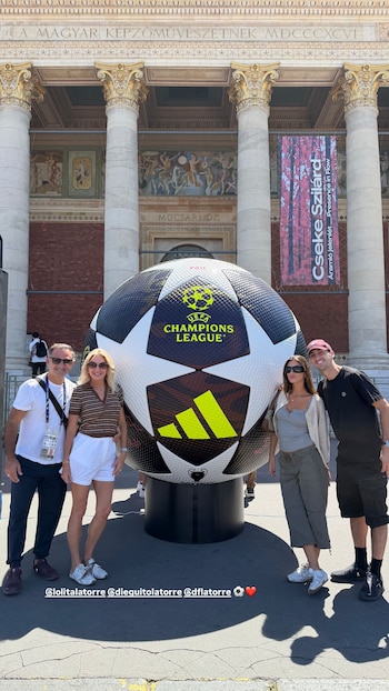 Cuatro personas, dos adultos y dos jóvenes, posan junto a un balón gigante de la Champions League frente a un edificio con columnas y murales en Budapest