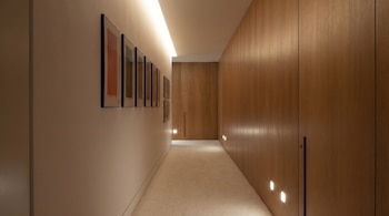Un largo pasillo con una pared beige que exhibe varias obras de arte enmarcadas y una tira de luz LED en el techo. La pared opuesta está cubierta con paneles de madera y luces de suelo.