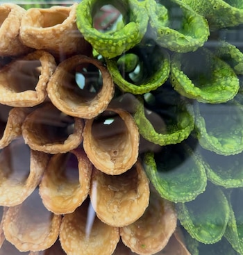 Primer plano de numerosas cáscaras de cannoli vacías; algunas son de color beige claro y otras de un verde brillante, dispuestas en una exhibición