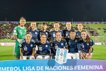Once jugadoras de la Selección Femenina de Argentina posan sonrientes en el campo de un estadio por la noche, vistiendo uniformes oscuros con detalles celestes y blancos